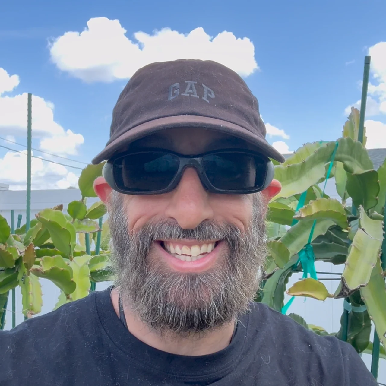 Jason Gentile, the Father of Dragons, smiling in a cap and sunglasses in front of his dragon fruit plants on their trellises.